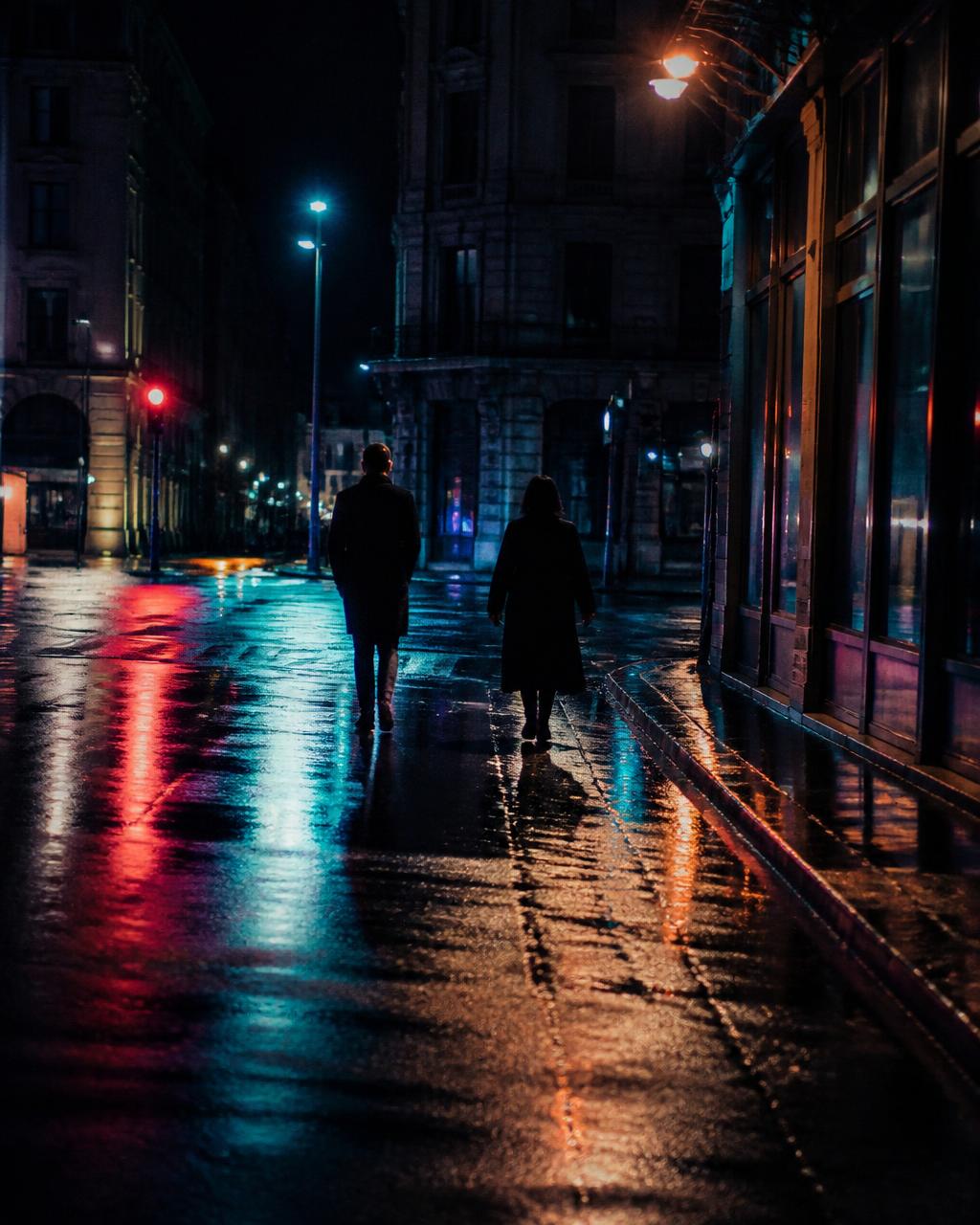 Two figures walking a rain-slick city street at night